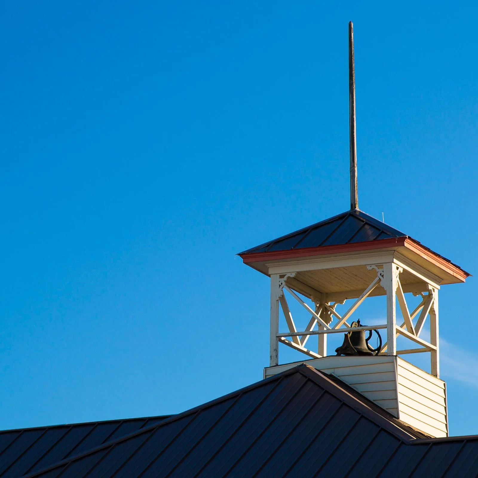 A white clock tower with a black bell inside and a red roof, set against a clear blue sky.