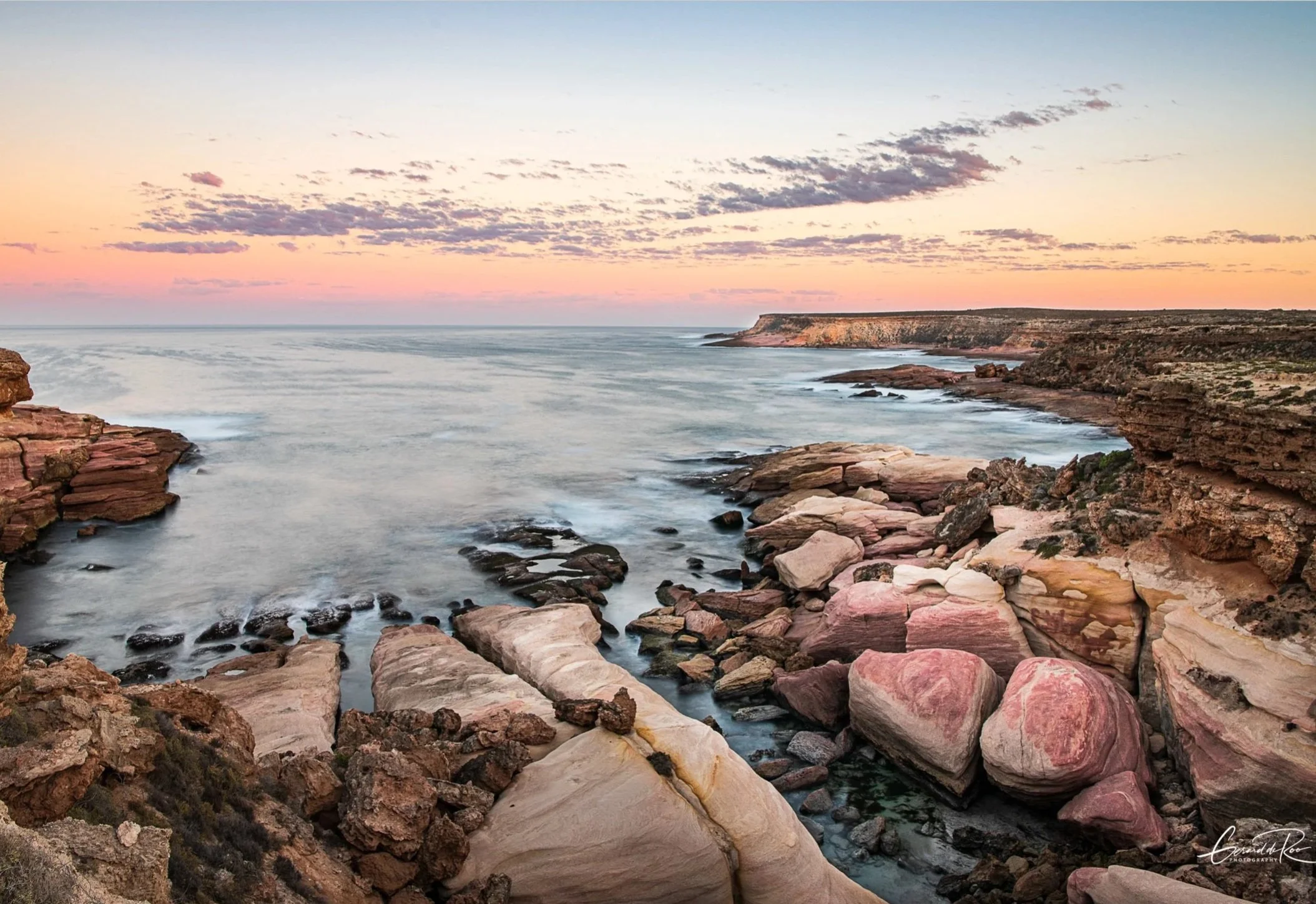 Sunset over rocky coastline with cliffs and pinkish rocks along the shore.