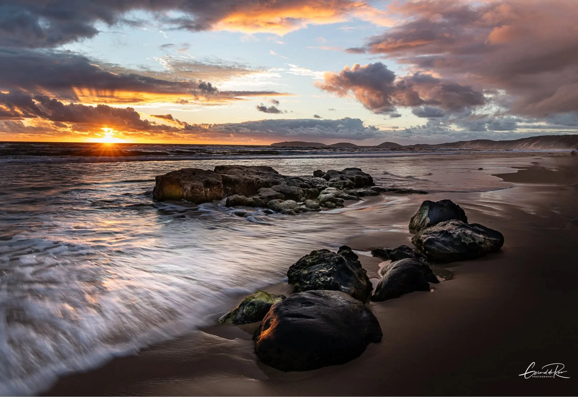 Sunset over a rocky beach with waves crashing, dark clouds in the sky, and a distant hilly landscape.