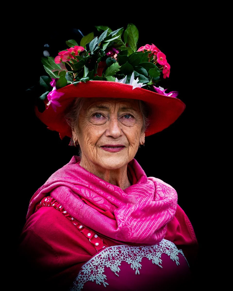 A senior woman wearing a large red hat decorated with green leaves and pink flowers, a pink scarf, and a pink shawl with lace details against a black background.