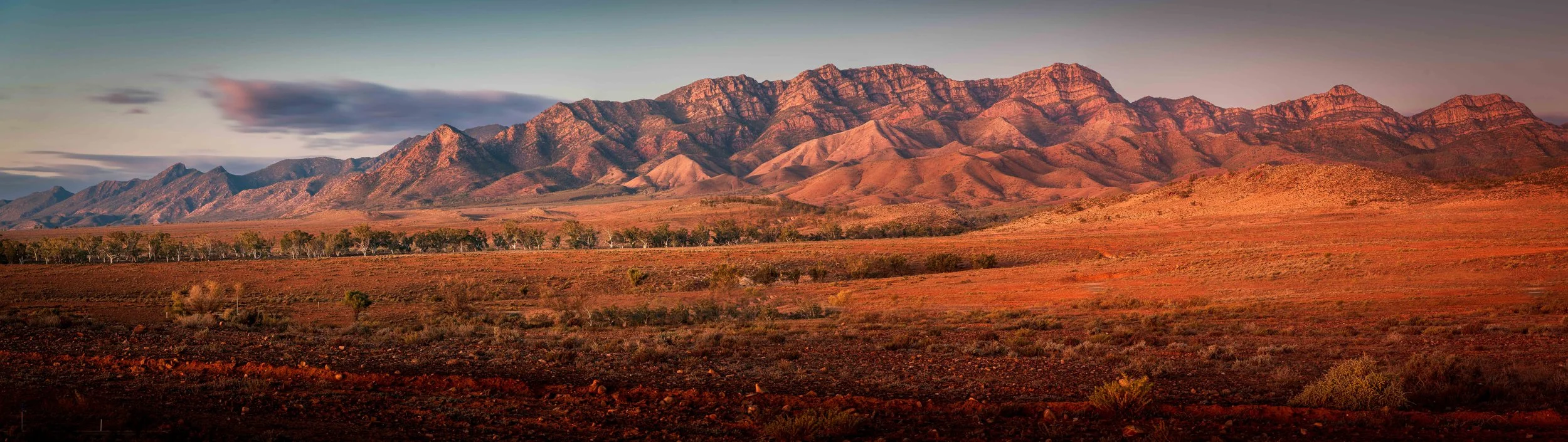 Sunset over a mountain range with red and orange hues, desert landscape, sparse vegetation, and a few clouds in the sky.