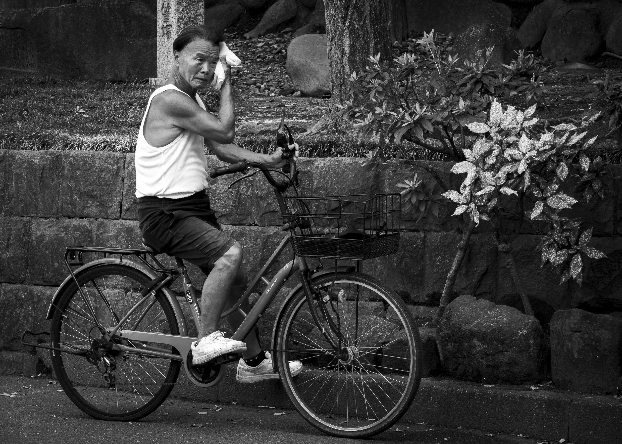 An elderly woman wearing a white tank top and dark shorts riding a bicycle with a basket on the front, wiping sweat from her forehead in an outdoor setting with large rocks and trees.