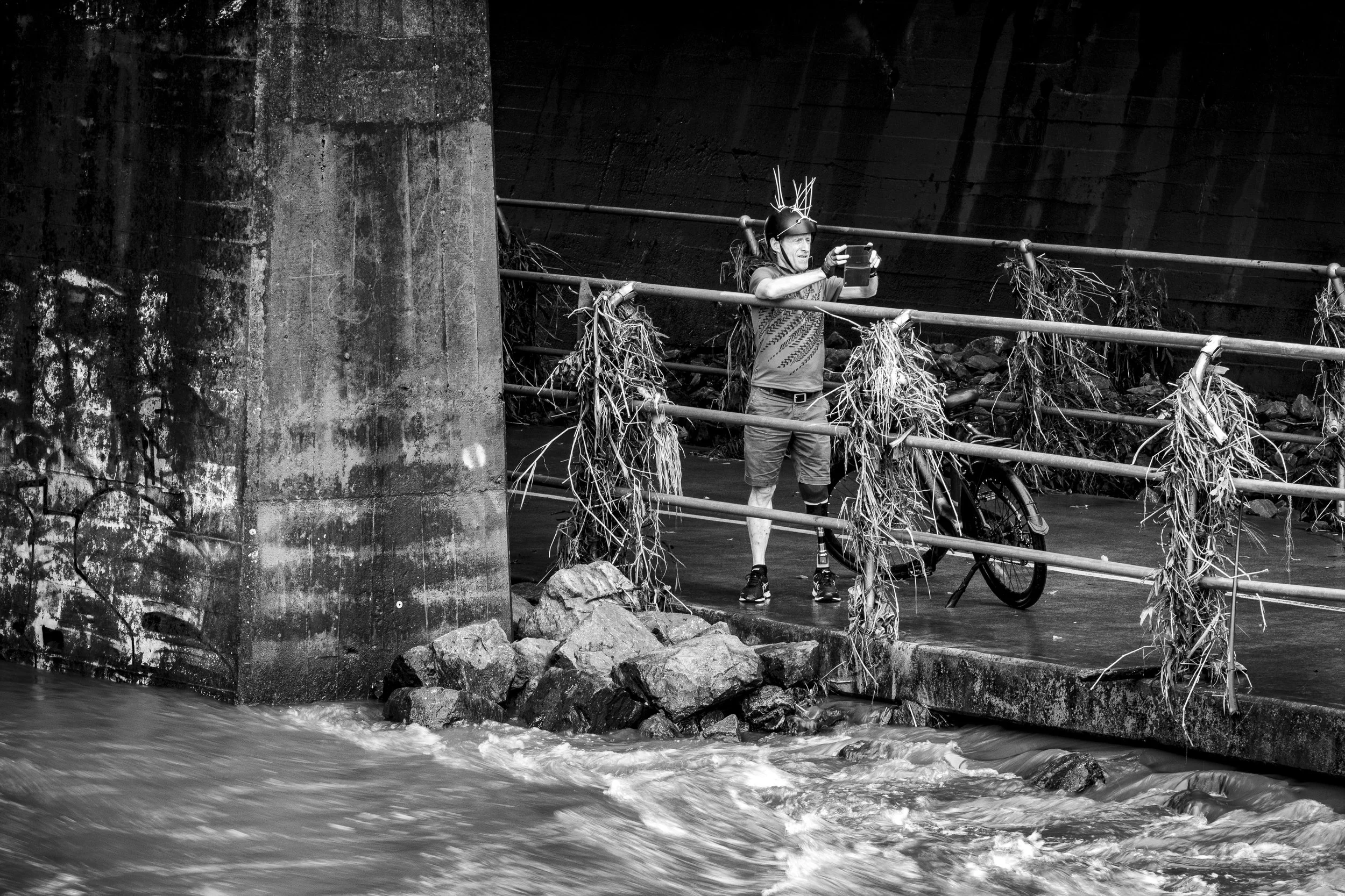 A person with a prosthetic leg taking a selfie with a smartphone on a bridge with water below, surrounded by debris and rocks.