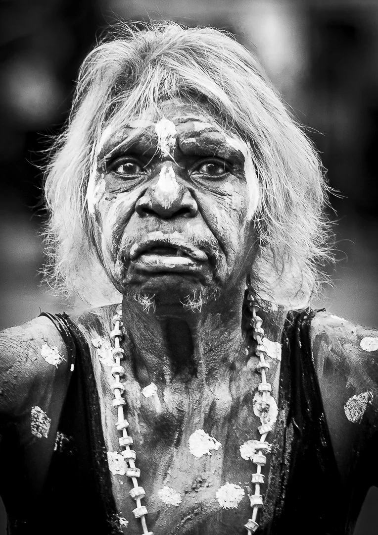 A black and white photo of an elderly indigenous woman with long, unkempt hair. She has a serious expression, with face paint or markings, and wears a necklace with large beads. Her face and body are decorated with white paint or markings.
