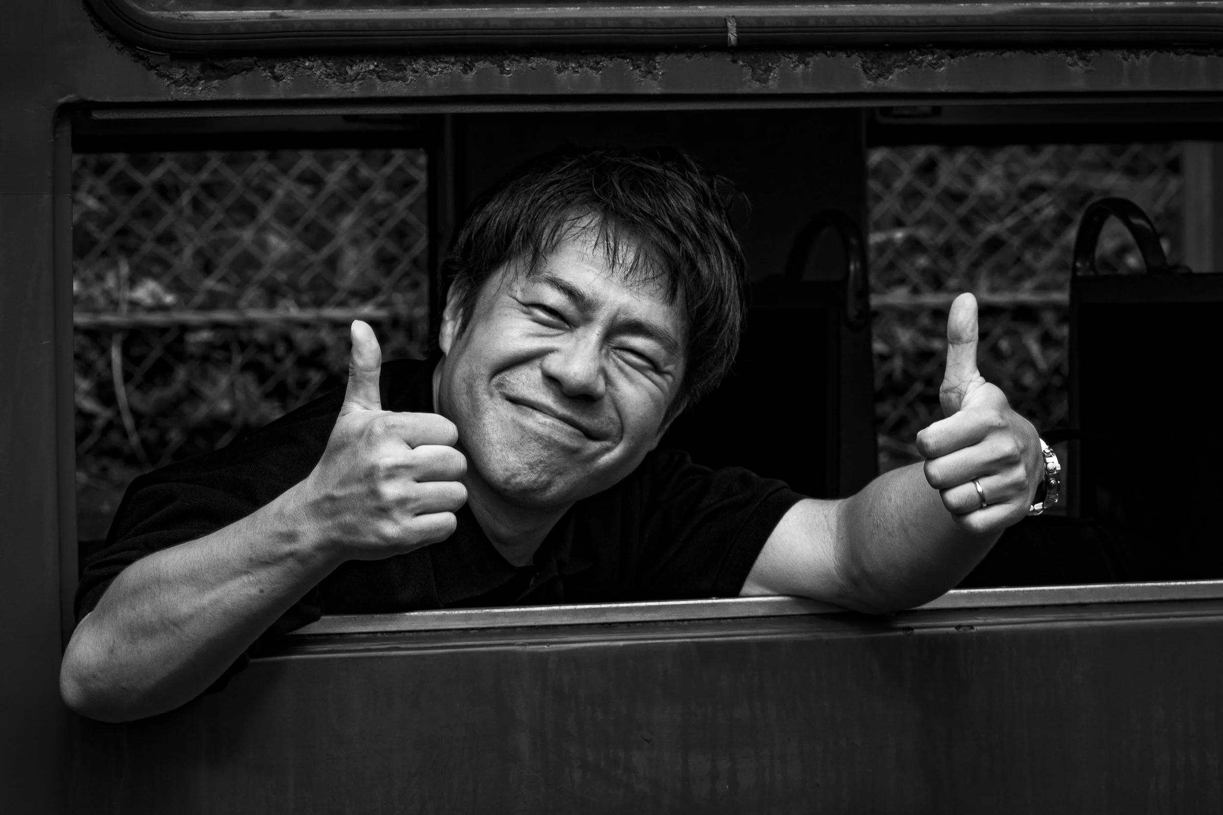 A man smiling and giving a thumbs-up gesture from a window, in black and white.