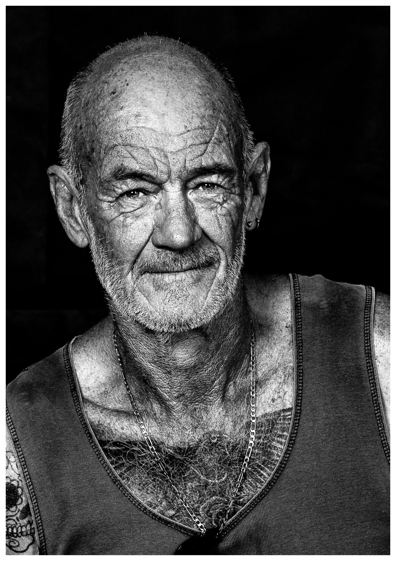 Close-up black and white portrait of an elderly man with a beard, tattoos, wearing a sleeveless shirt, and a chain necklace.