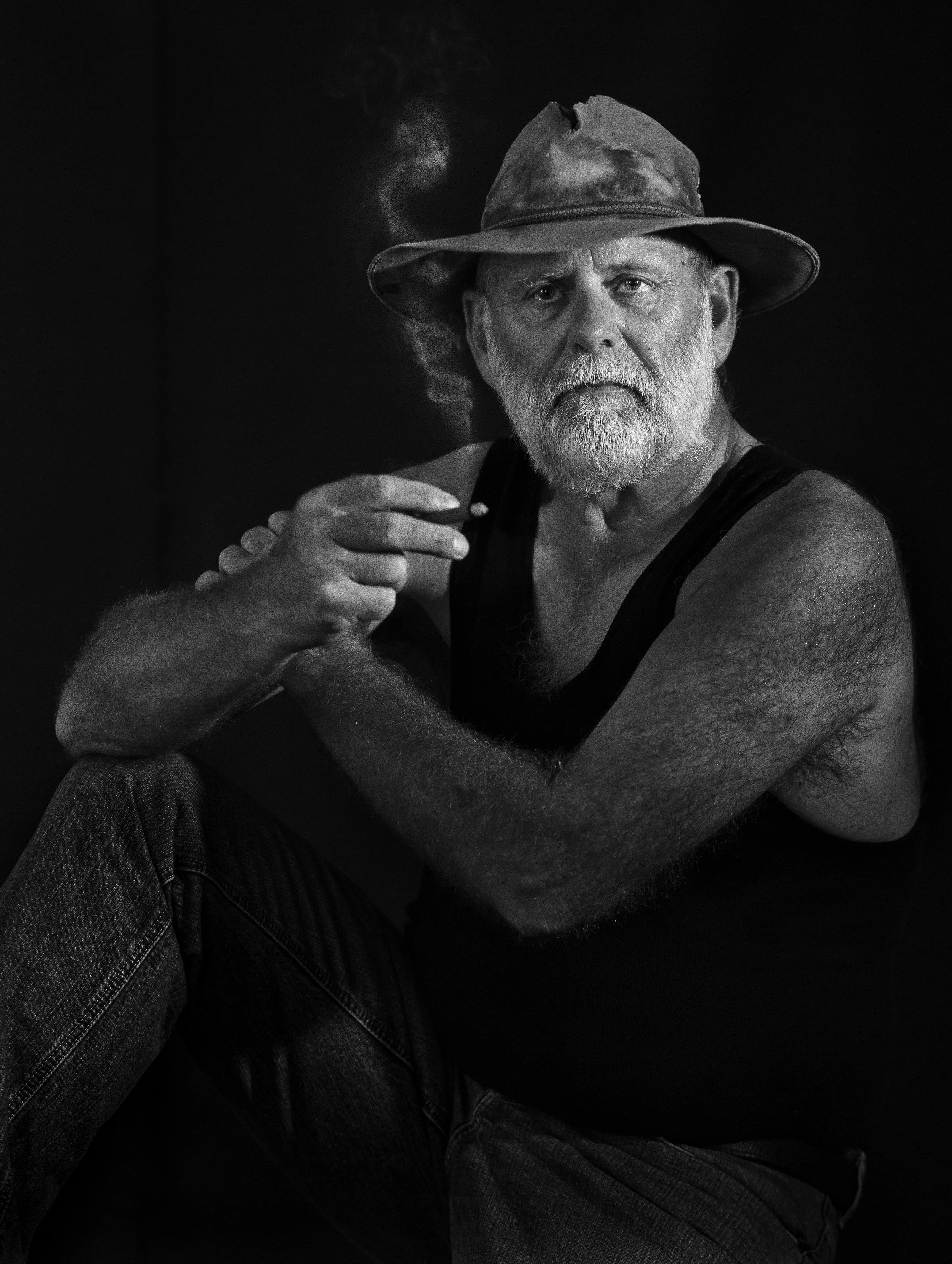 Black and white photo of an older man with a beard wearing a hat and a tank top, smoking a cigarette, with a serious expression.