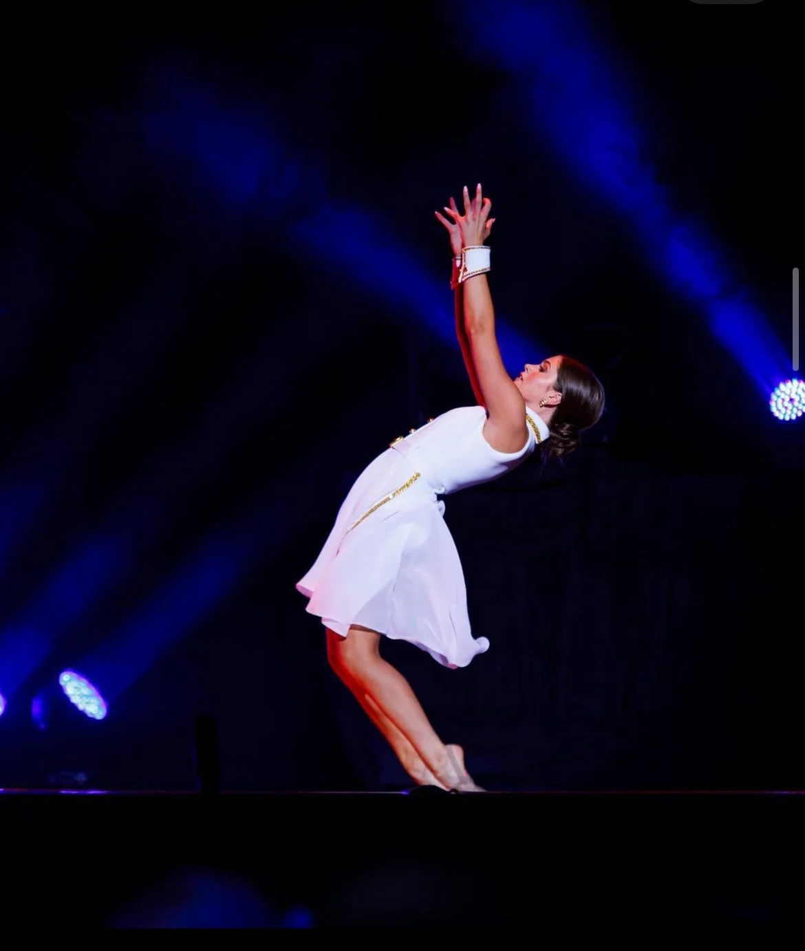 A woman performing on stage, wearing a white dress, with arms raised, in front of a dark background with blue stage lights.