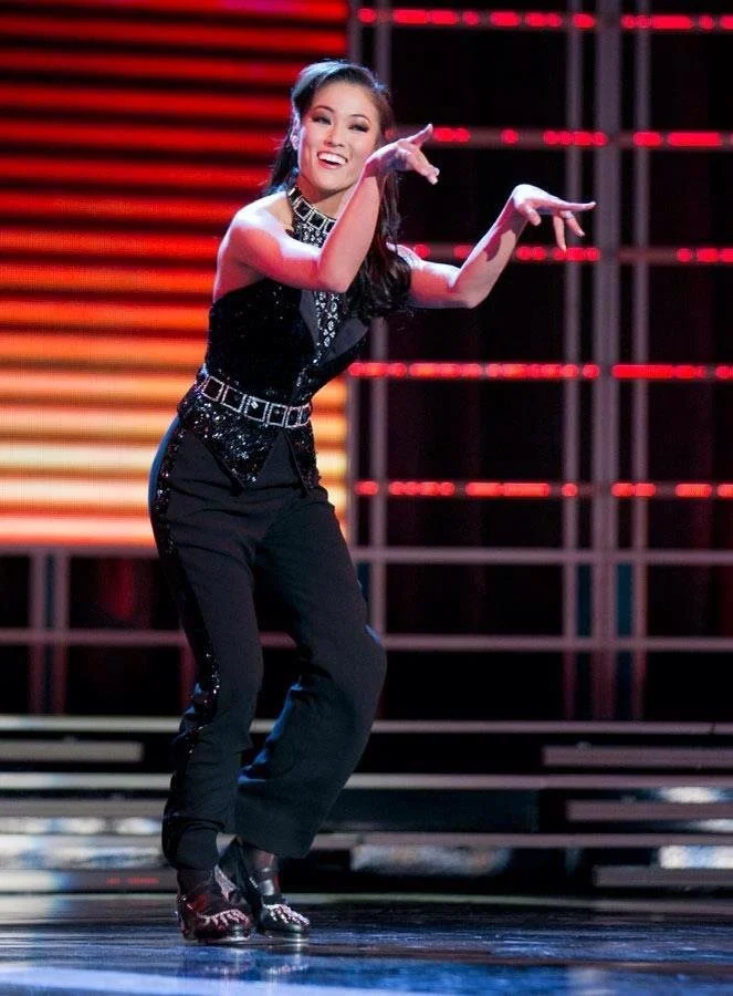 A woman dressed in a black sequined outfit dancing on a stage with colorful background lights.