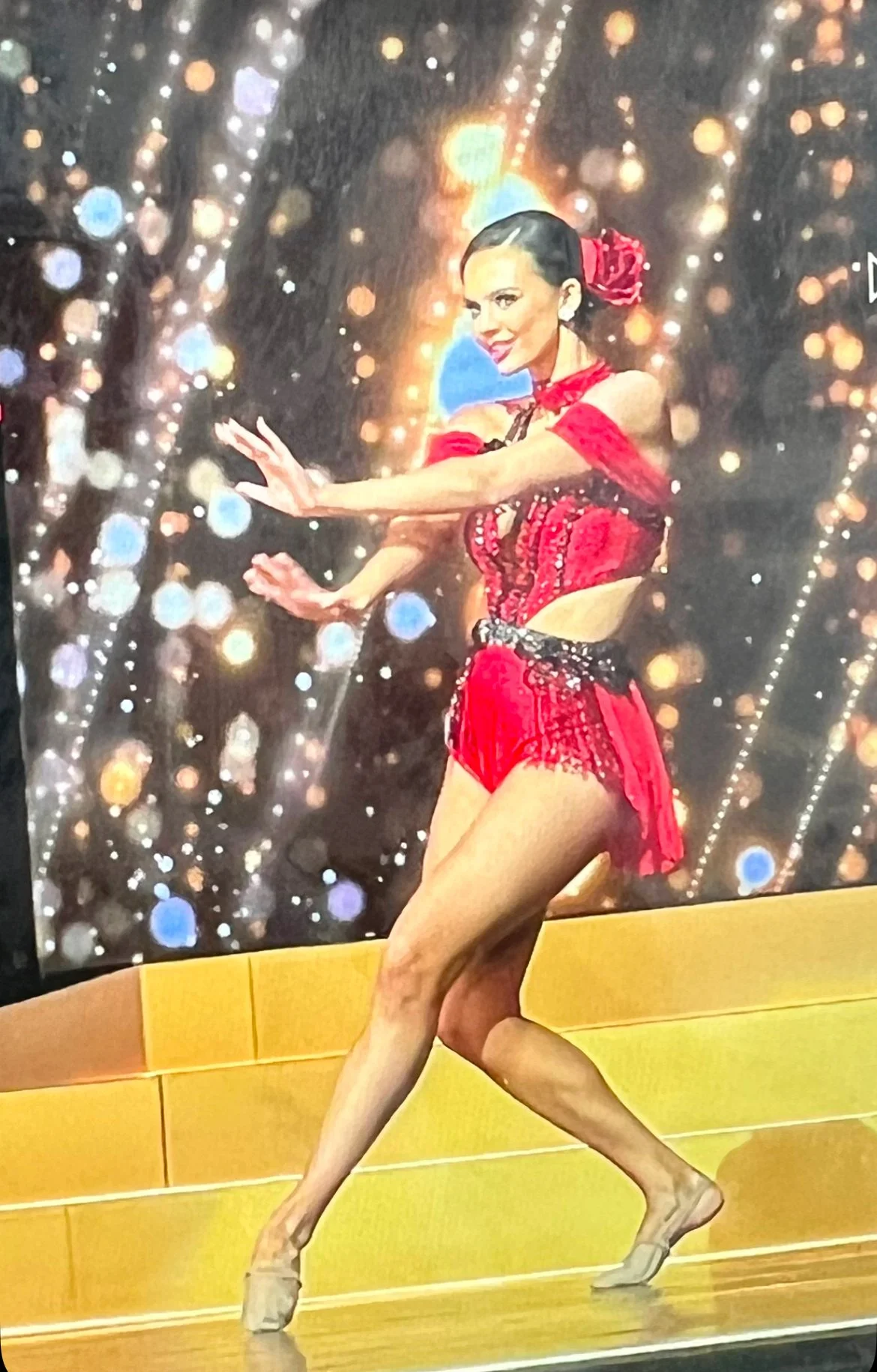A woman in a red and black ballroom dance costume poses on a stage with a sparkling, bokeh background.