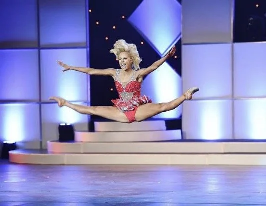 A woman with blonde hair jumping in the air with her legs split and arms outstretched on a stage with blue panels in the background.