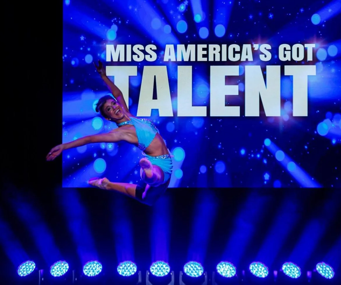 A female performer in a blue costume is jumping in front of a large screen that reads 'Miss America's Got Talent' with a blue sparkling background.