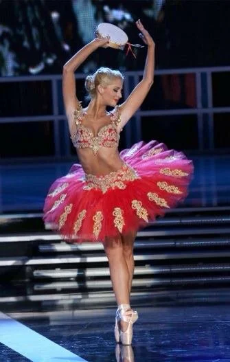 Ballerina in a pink tutu with gold embellishments, performing a dance on stage, holding a white fan above her head.