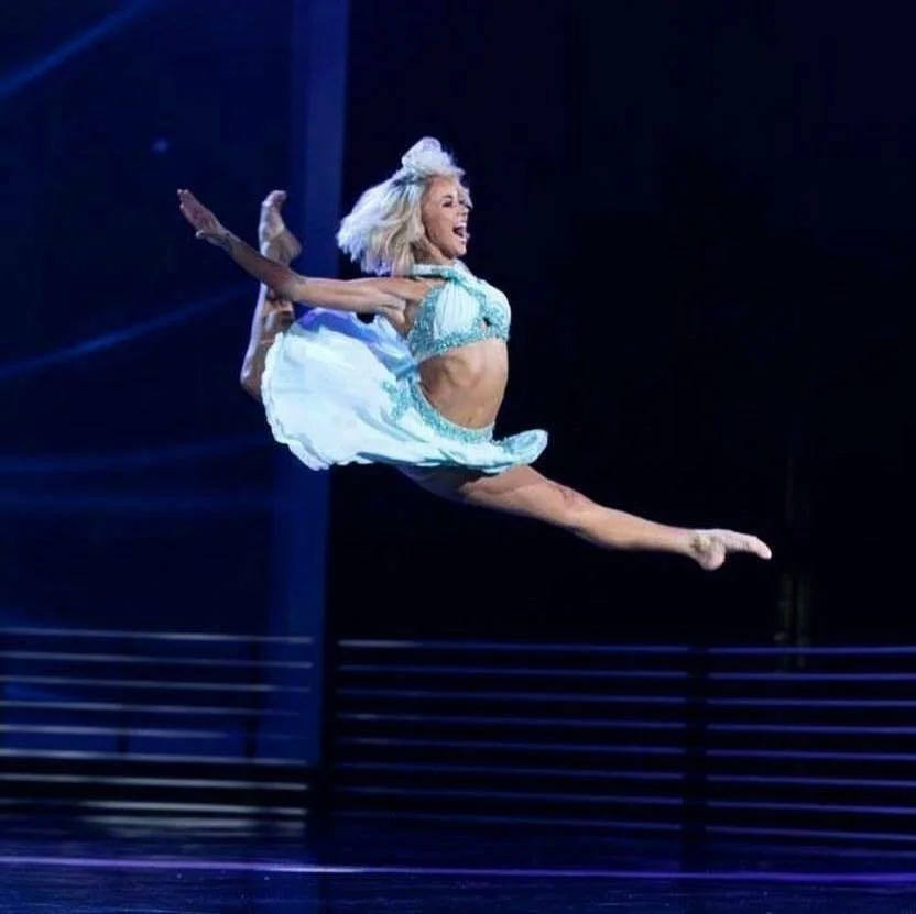 A woman in a light blue costume performing a dance leap on stage.