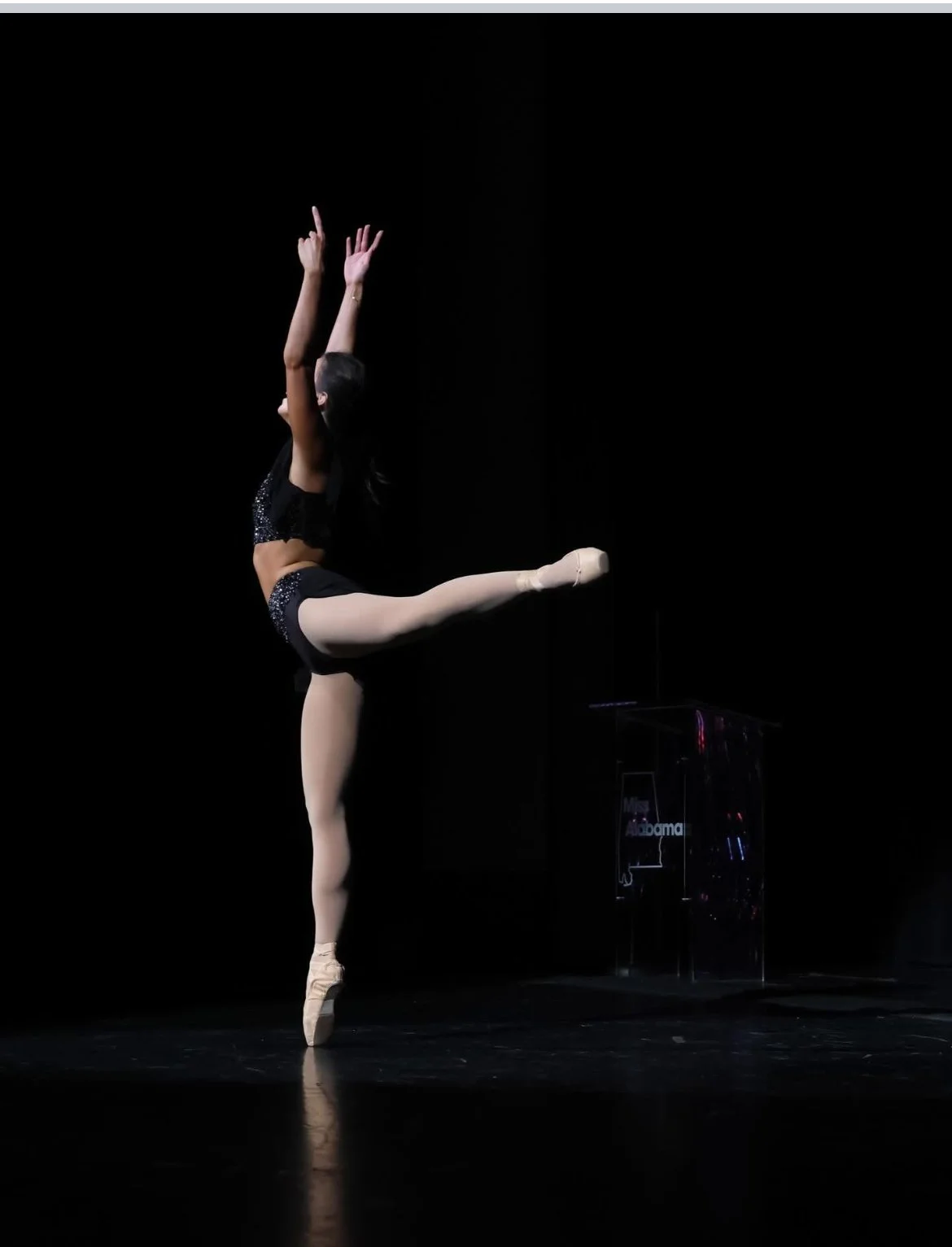 A ballet dancer performing on stage in black costume, standing on one leg with arms raised, against a dark background with a podium labeled 'Miss Alabama'.