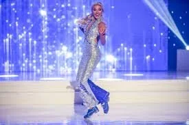 A woman in a shiny silver outfit and ice skates performing on an ice rink stage with blue lighting and sparkles.