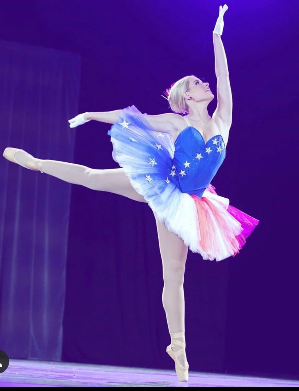 Ballet dancer in a blue star-themed tutu performs an arabesque on stage with purple lighting.