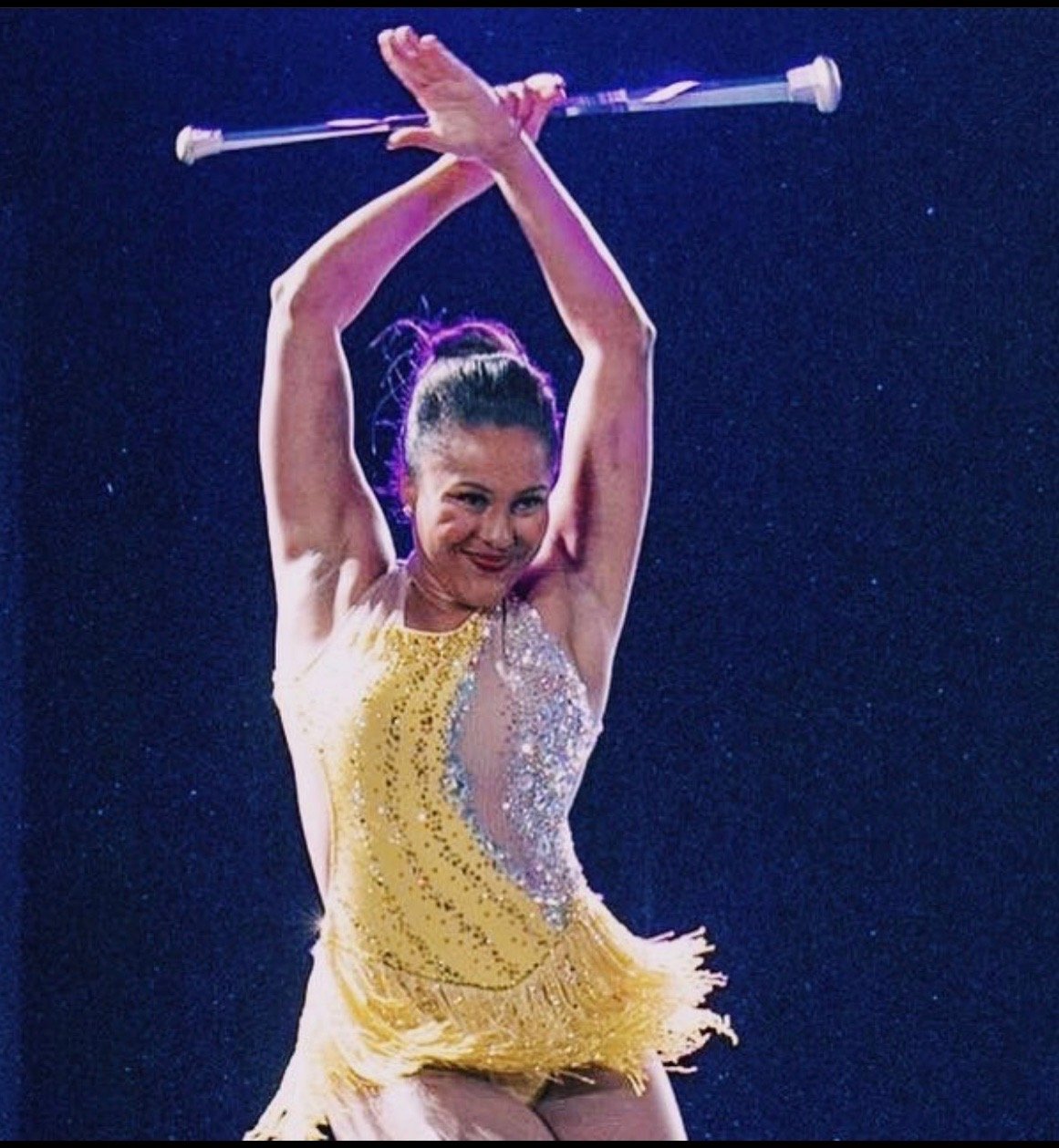 A female gymnast dressed in a yellow leotard with rhinestones, performing a routine with a javelin against a dark background.
