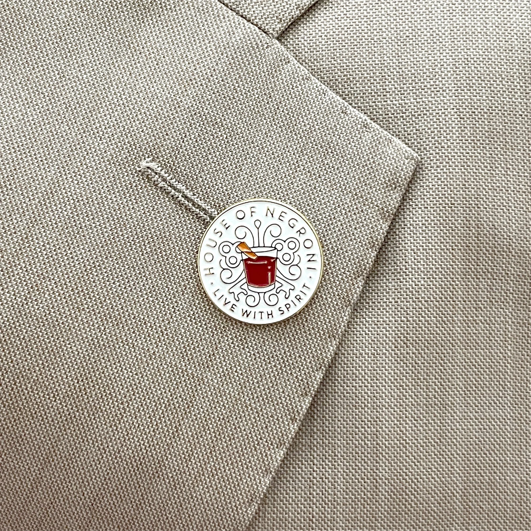 A lapel pin with the logo of the House of Negroni, featuring a red cocktail glass and the words 'House of Negroni' and 'Live with Spirit' on a beige fabric suit lapel.
