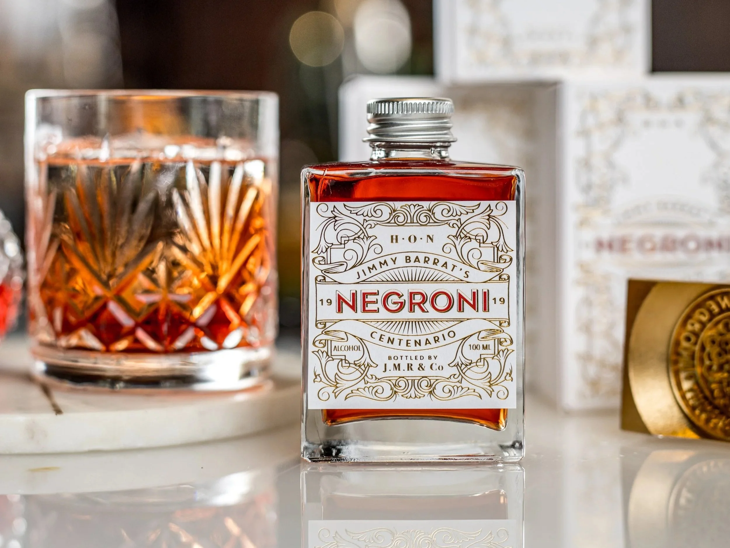 A bottle of Negroni with an ornate label design on the front of the bottle. To the left of the image is a negroni cocktail served in a glass.