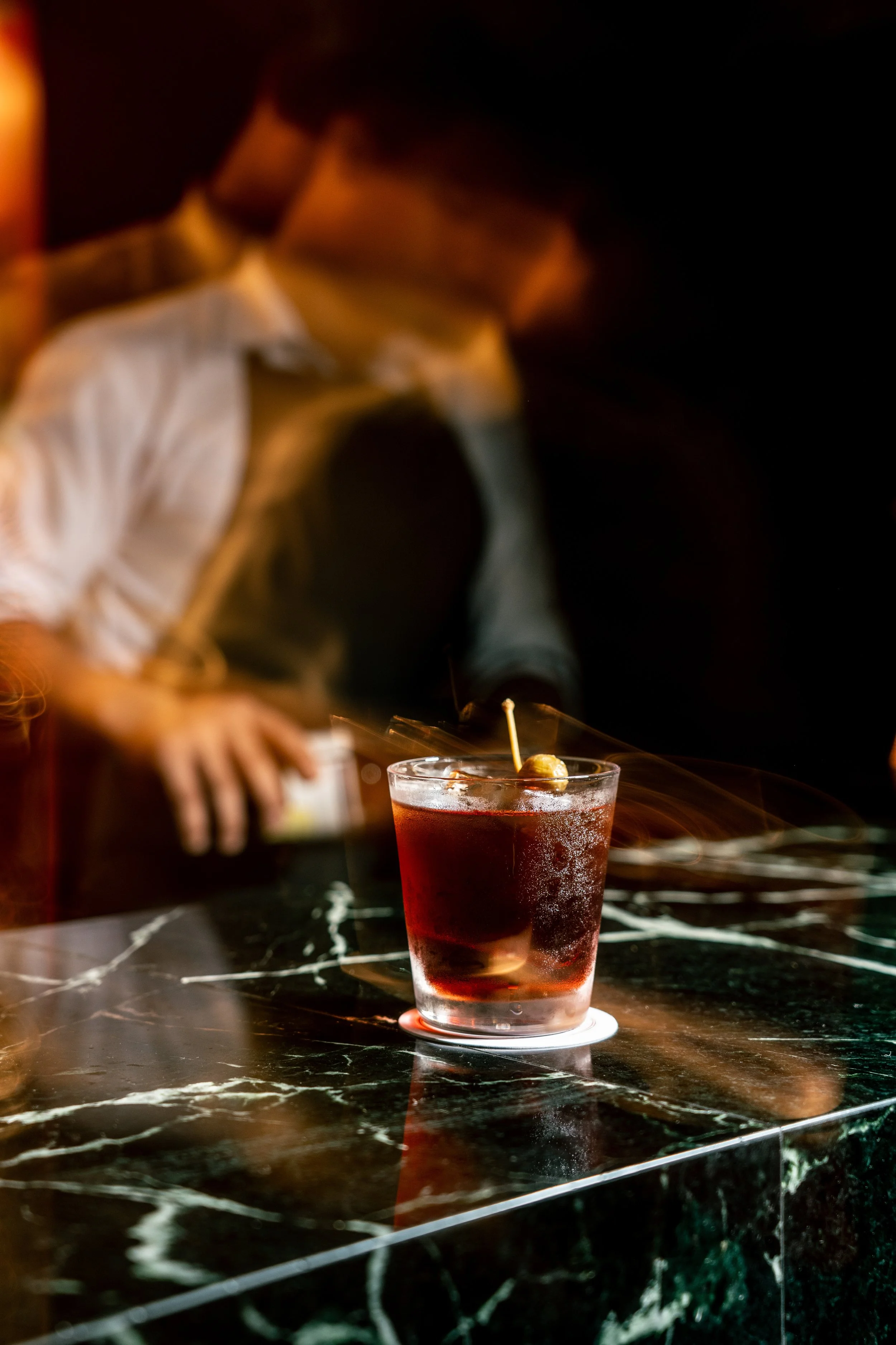 A rocks glass with a negroni cocktail garnished with a green caper, sitting on a black marble bar counter with white veins; a blurred bartender in the background.