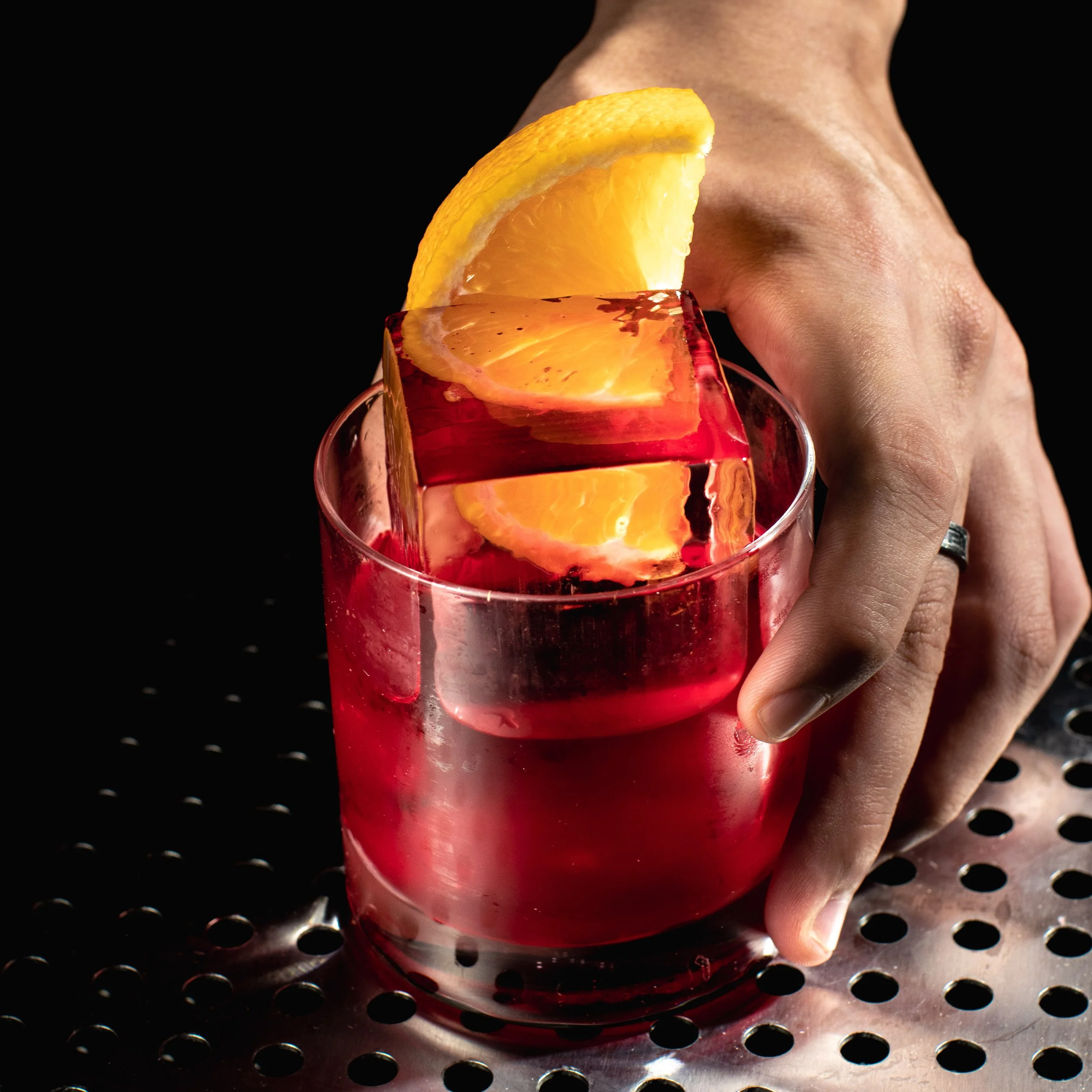 A hand holding a transparent glass filled with a red Negroni cocktail, garnished with orange slices, on a metallic surface with black background.