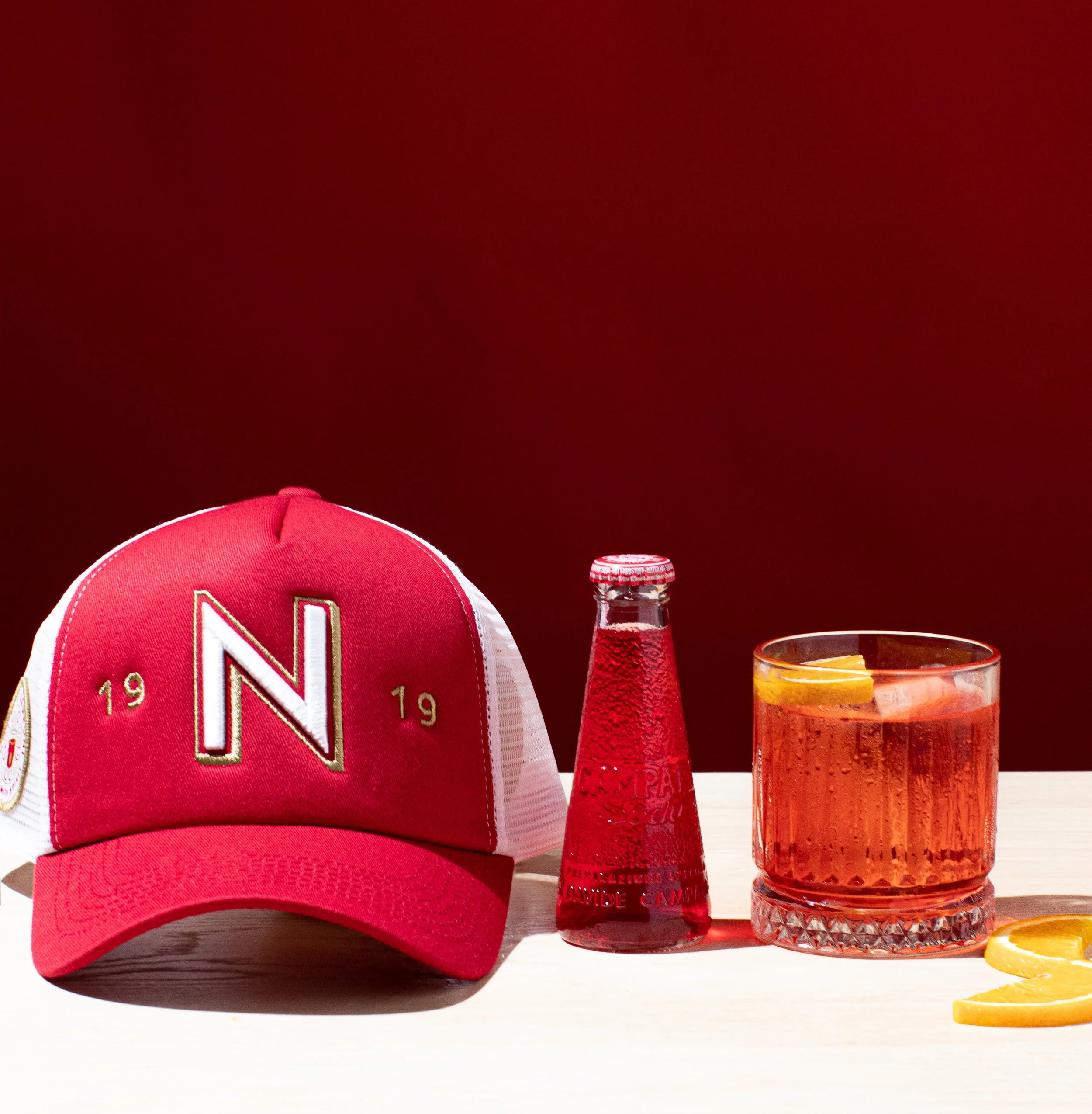 Red and white trucker hat with a large 'N' logo, a glass bottle of red Campari soda, a glass of Negroni with orange slices, and an orange slice on a light-colored surface against a dark red background.