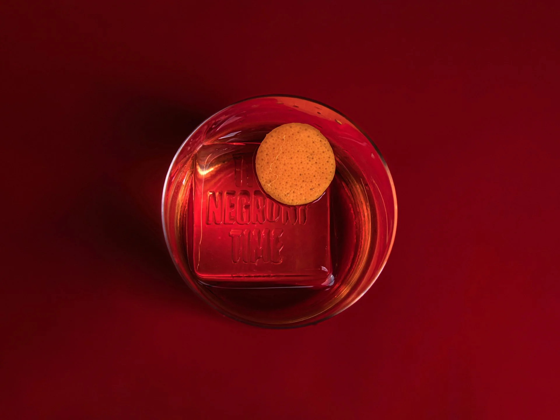 Top-down view of a glass with red-colored liquid and a round yellow orange peel sitting on the rim, with ice inside the glass. The ice is embossed to read 'NEGRONI TIME'. The image is on a red background.