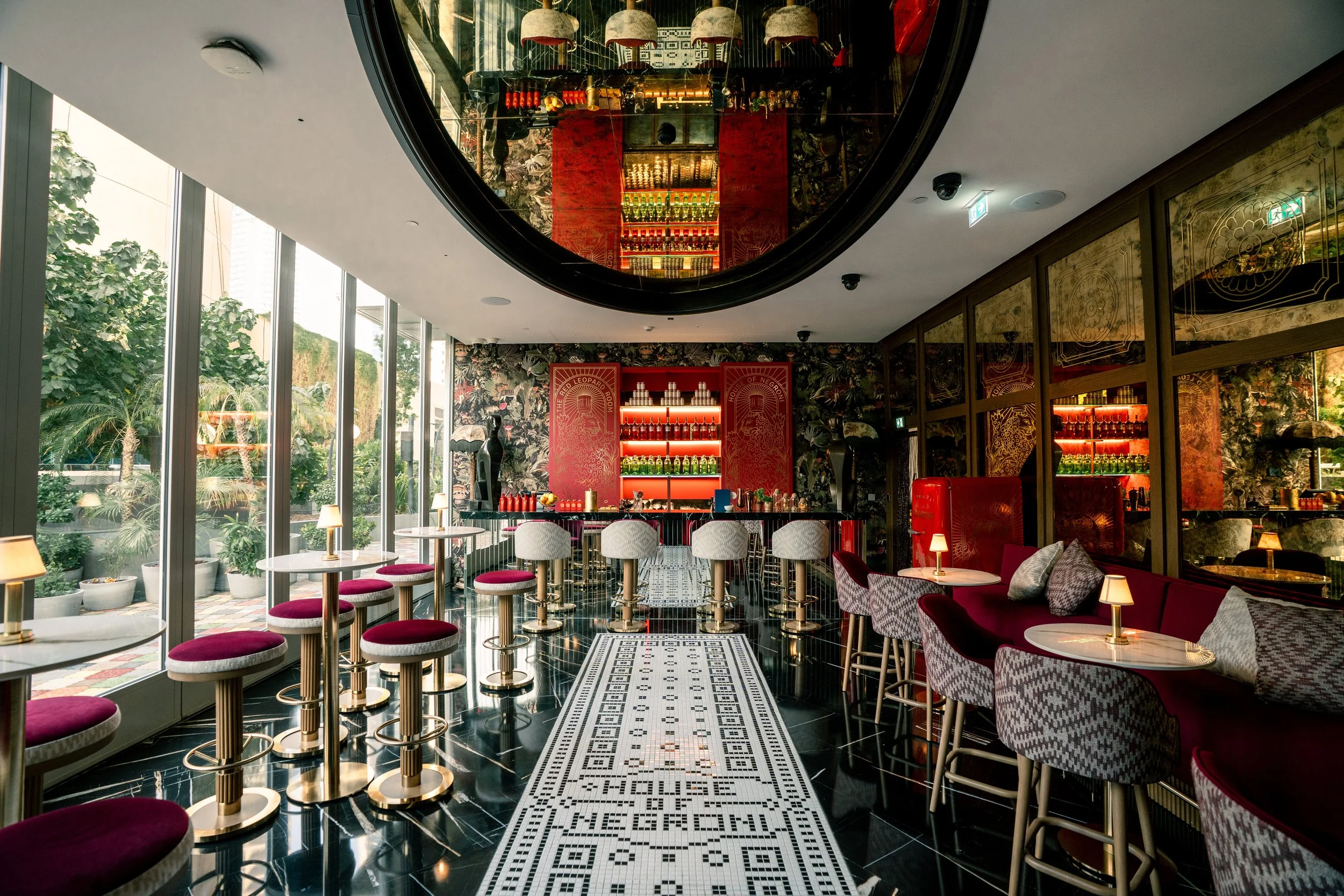 Stylish bar with high-top tables and bar stools near large windows, ornate decor including wallpaper, shelving with bottles, and a mirrored ceiling reflecting the room. Red, white, and black color theme.