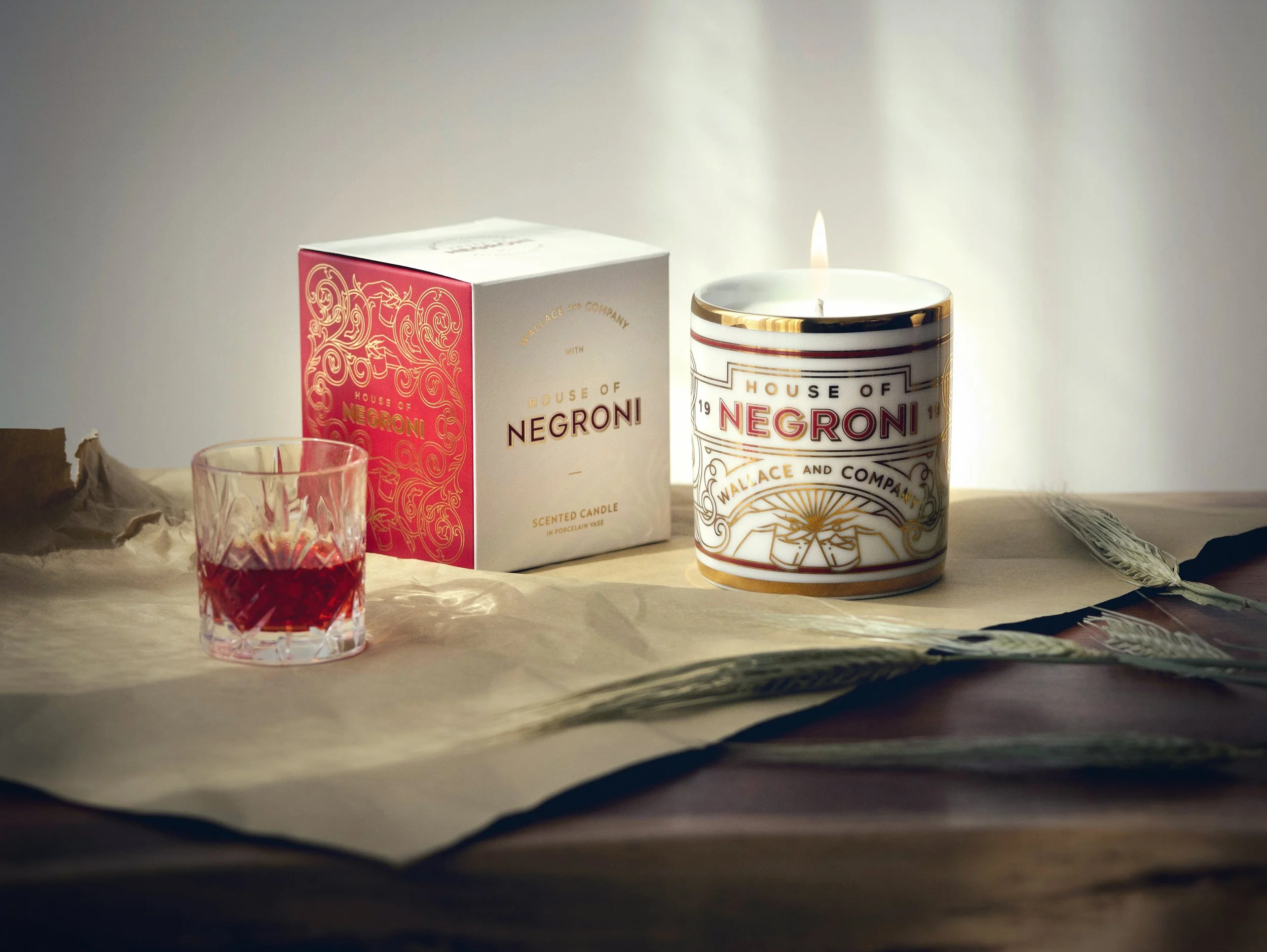 A decorative scented candle labeled 'House of Negroni' with a small glass containing red liquid and a white box with gold and red designs on a textured surface with dried wheat stalks.