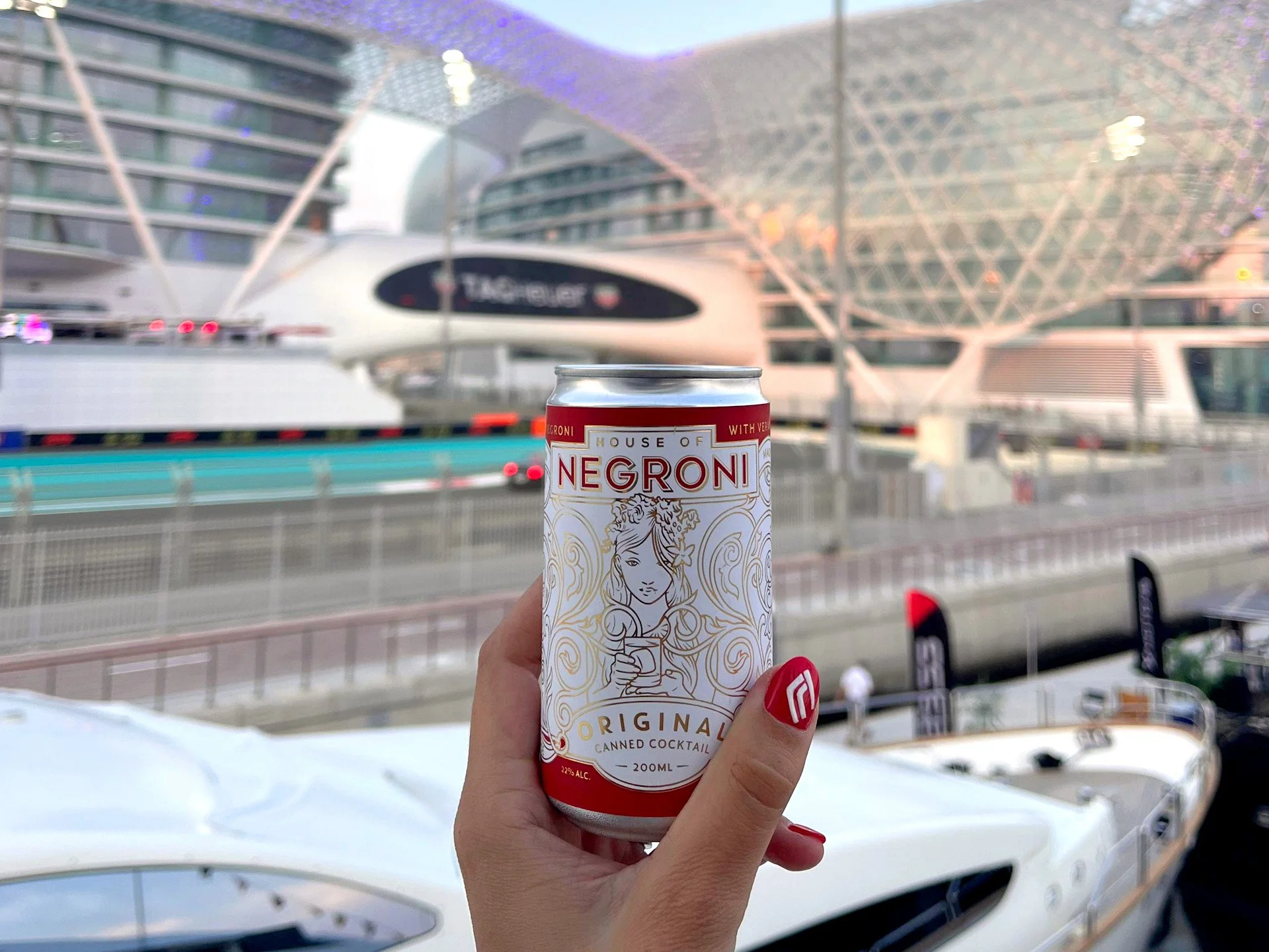 A female hand with red nails holding up a canned house of negroni cocktail, overlooking the Yas Marina circuit.