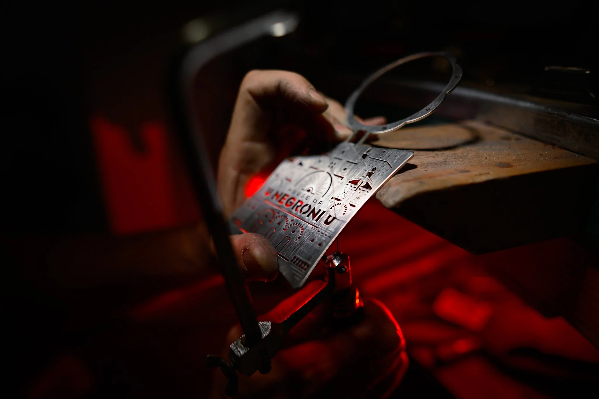 Close-up of a craftsman's hand holding a metal jewelry design template over a workbench, illuminated by a red light in a workshop.