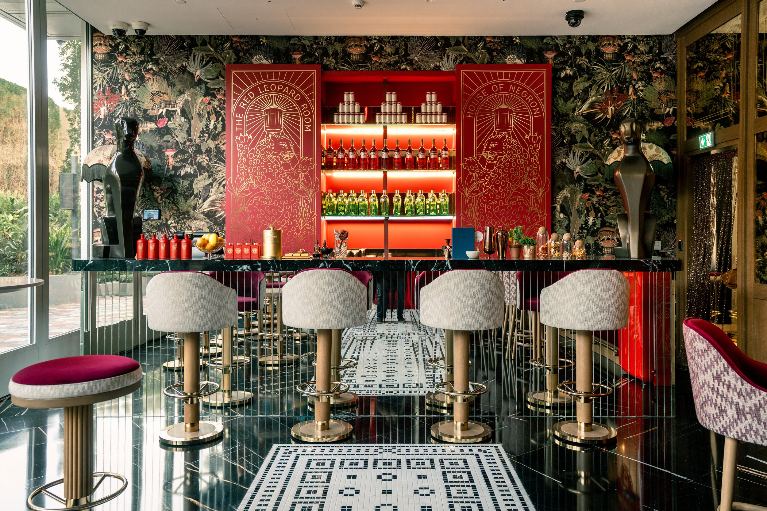 Interior of a modern bar with a black marble counter, featuring decorative red panels labeled 'The Red Leopard Room' and 'House of Negroni.' The background has a floral wallpaper with tropical plants. There are white and maroon bar stools around the counter, sculptures flanking the bar, and shelves with bottles and cans of alcohol. Large windows on the left let in natural light and display greenery outside.