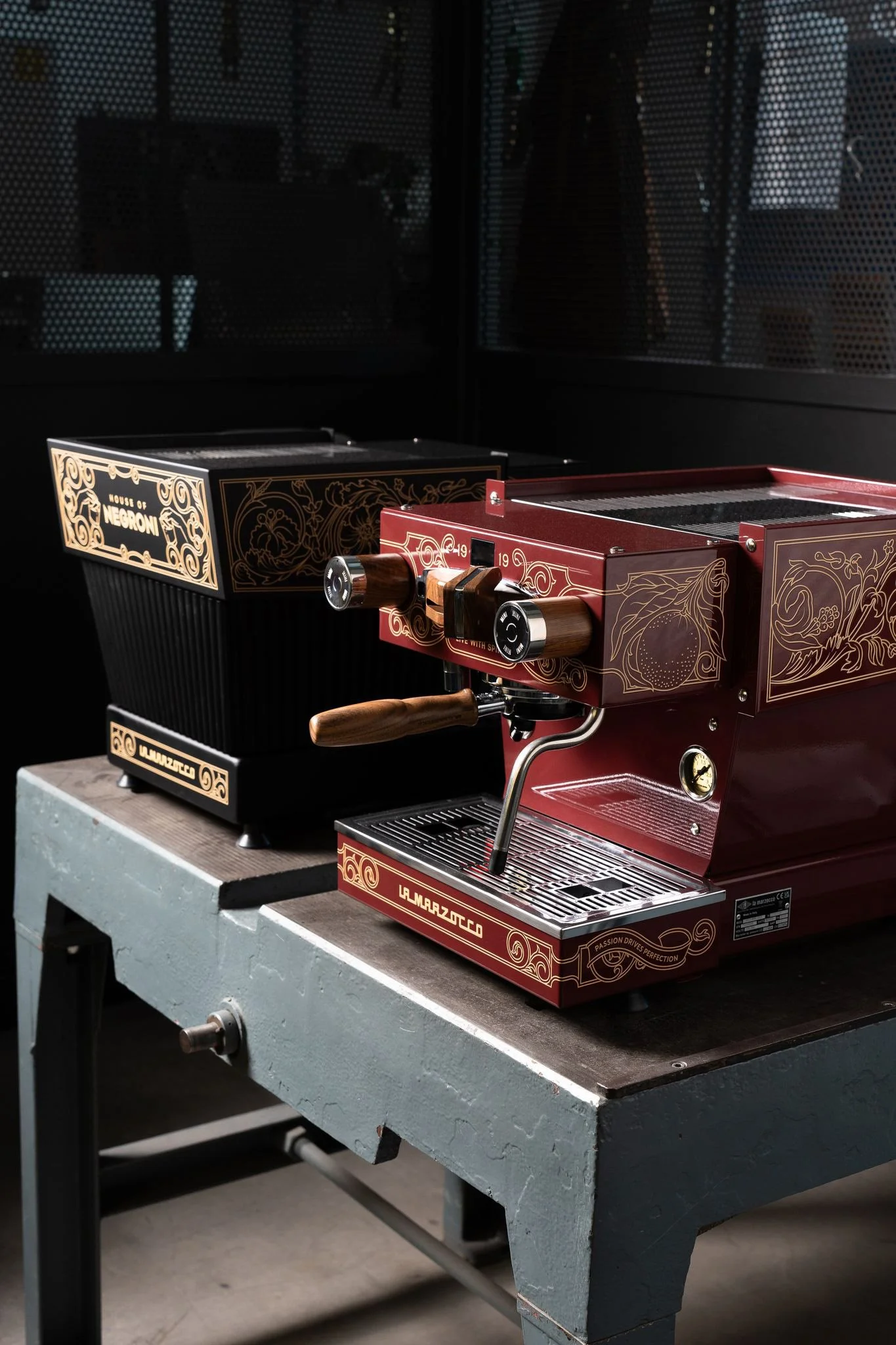 Two coffee machines next to each other, a burgundy one facing forward, a black one facing back. An elegant ornamental design adorns the facade of both machines.