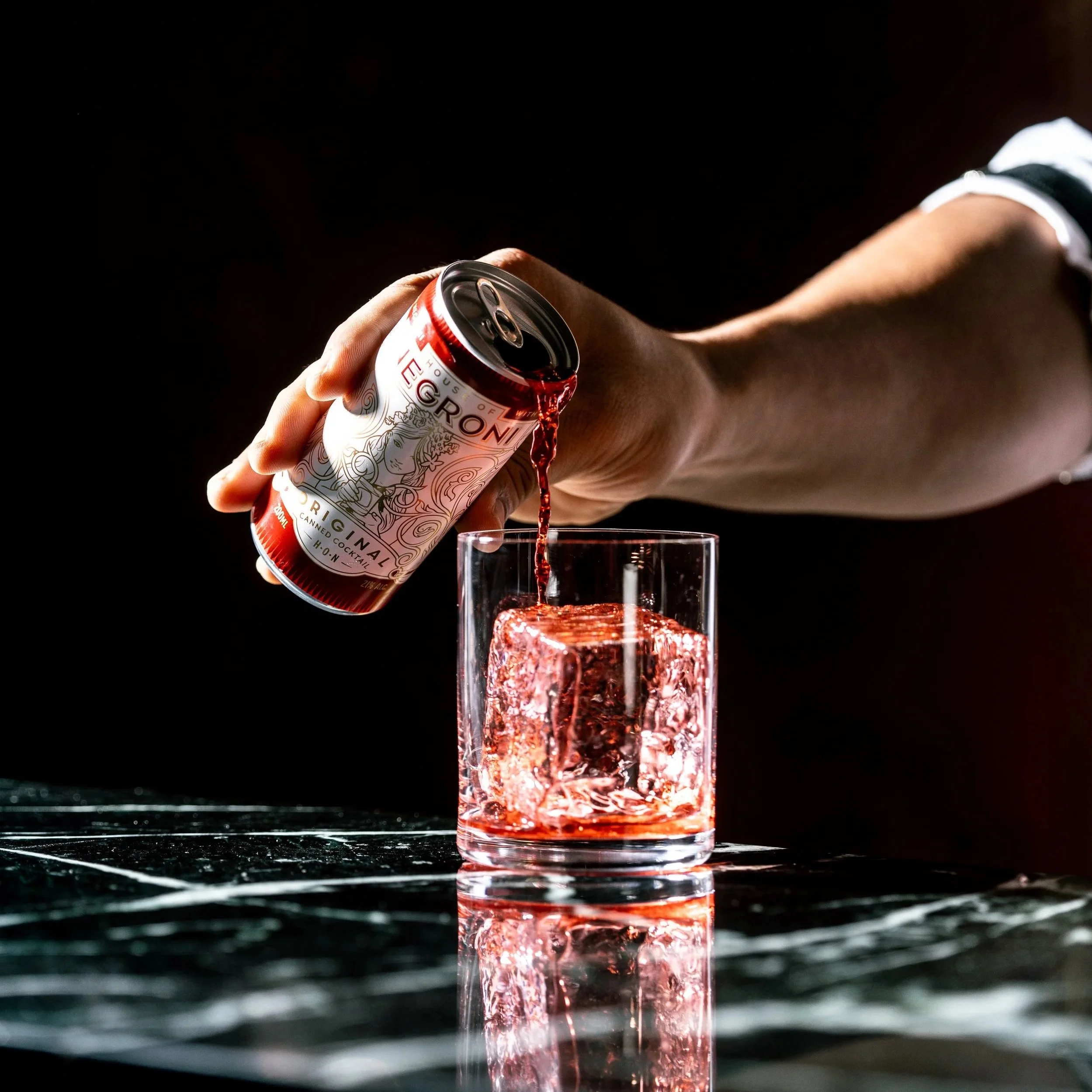 Person pouring a House of Negroni canned cocktail beverage from a can into a glass with a large ice cube on a dark, reflective surface