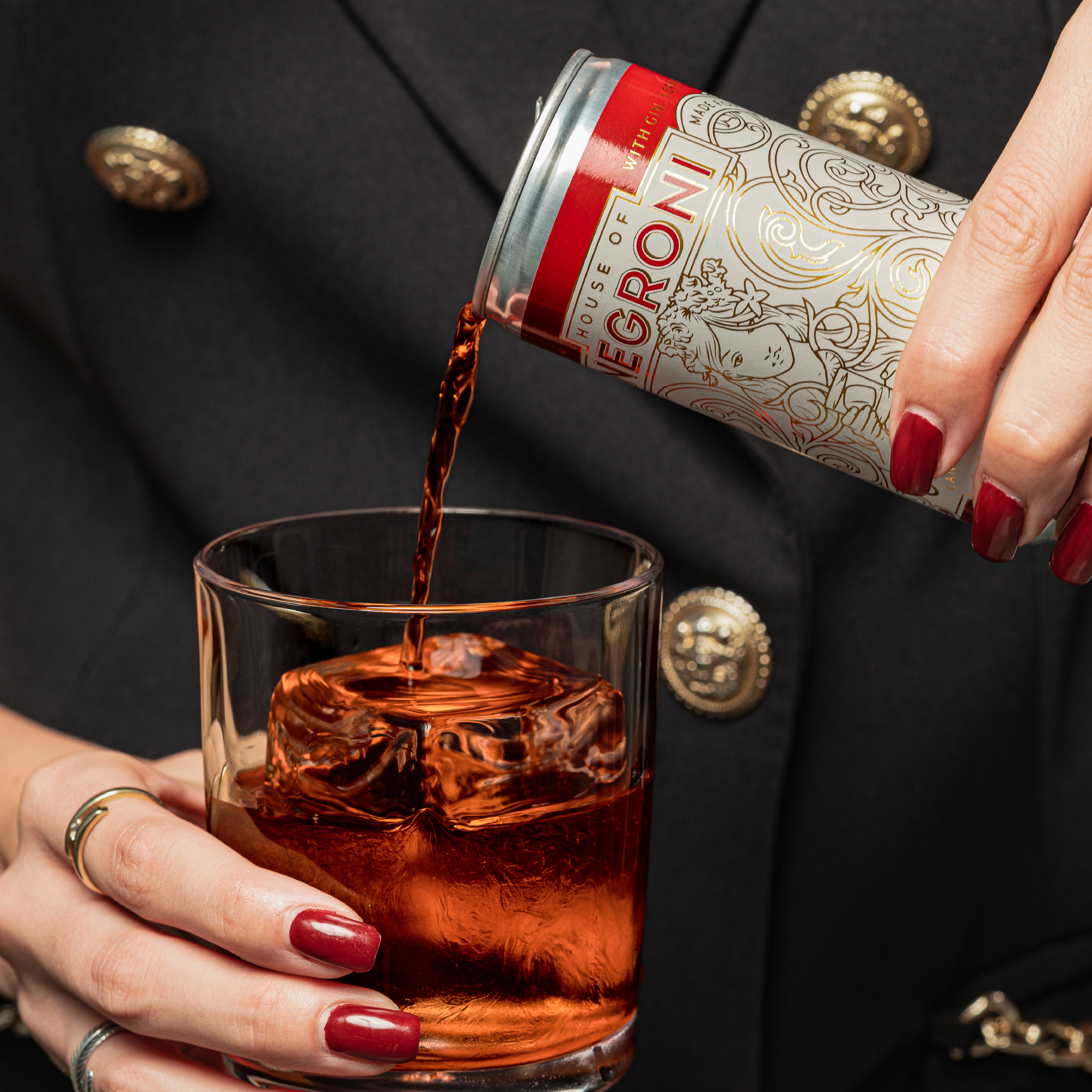 Person with red painted nails and gold rings pours a cocktail from a decorated can into a glass with a large ice cube.