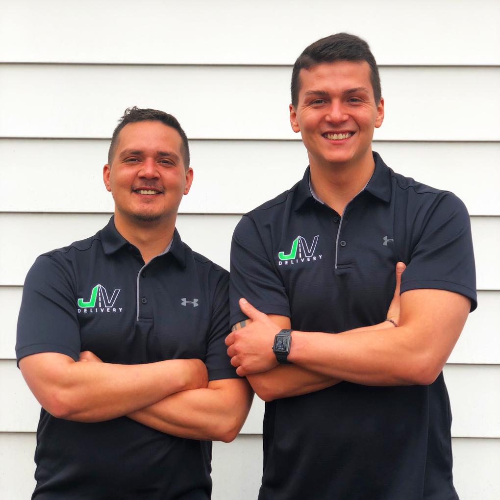 Two smiling men stand side by side with arms crossed, wearing black polo shirts with a logo that reads 'JV Delivery' on the chest, against a white horizontal siding wall.