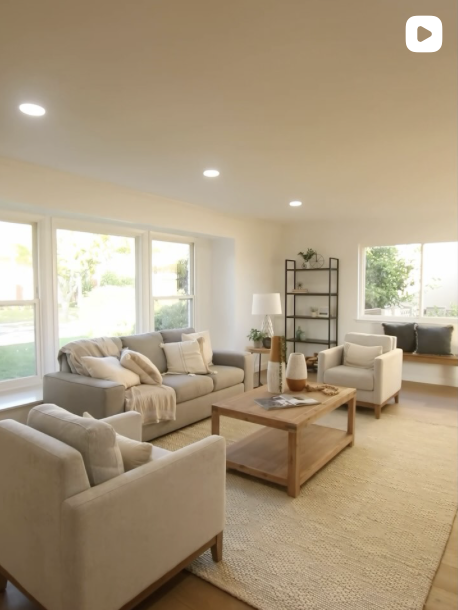 Bright living room with cream-colored sofas, a wooden coffee table, a black metal shelving unit, and large windows letting in natural light.