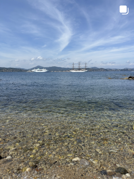Clear water with a rocky shoreline, sailboats and a cruise ship on the water, and mountains in the distance under a partly cloudy sky.