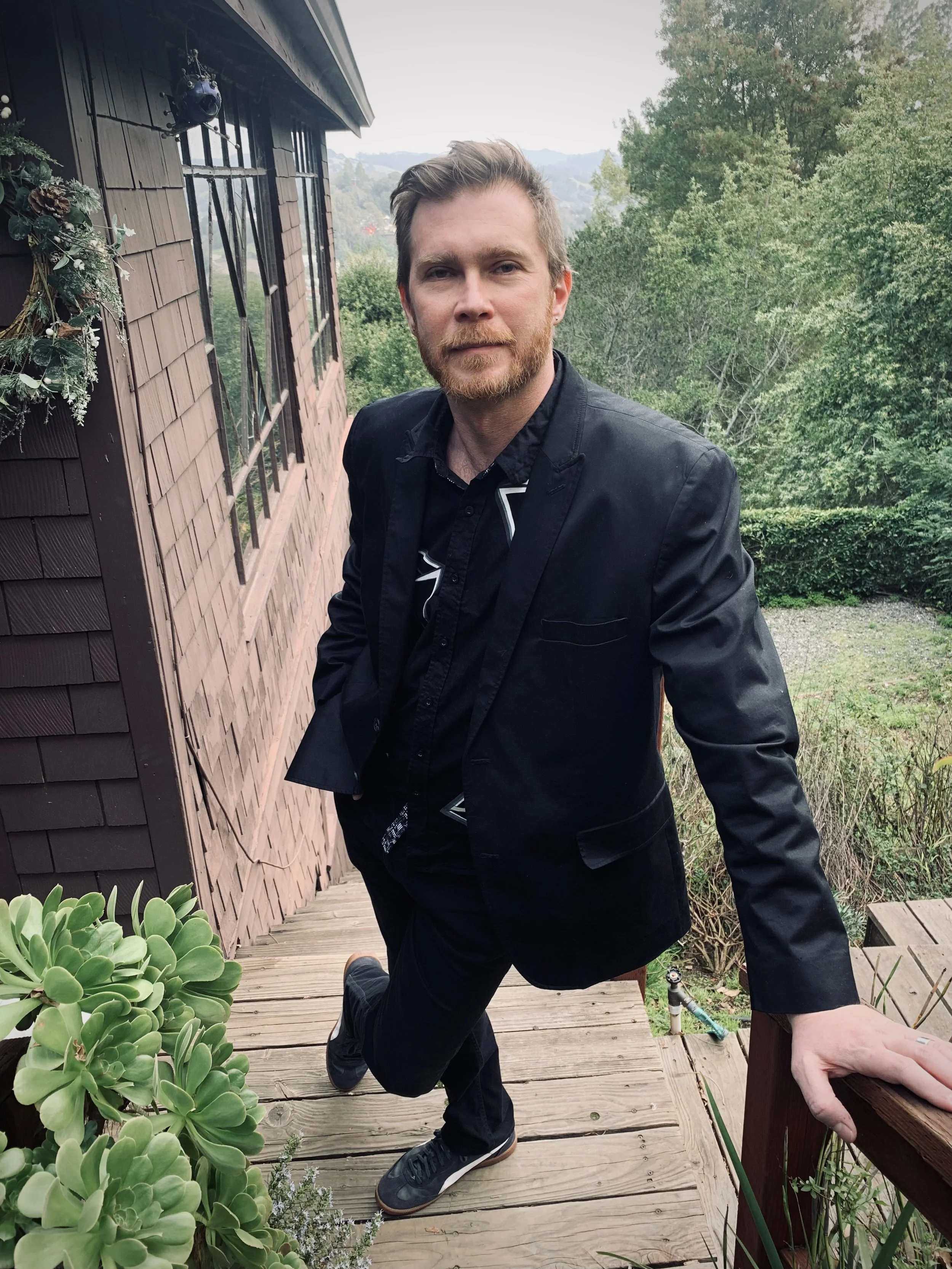 A man in a black suit and black sneakers leaning on a wooden railing outside a brown house with shingle siding, surrounded by greenery and trees.