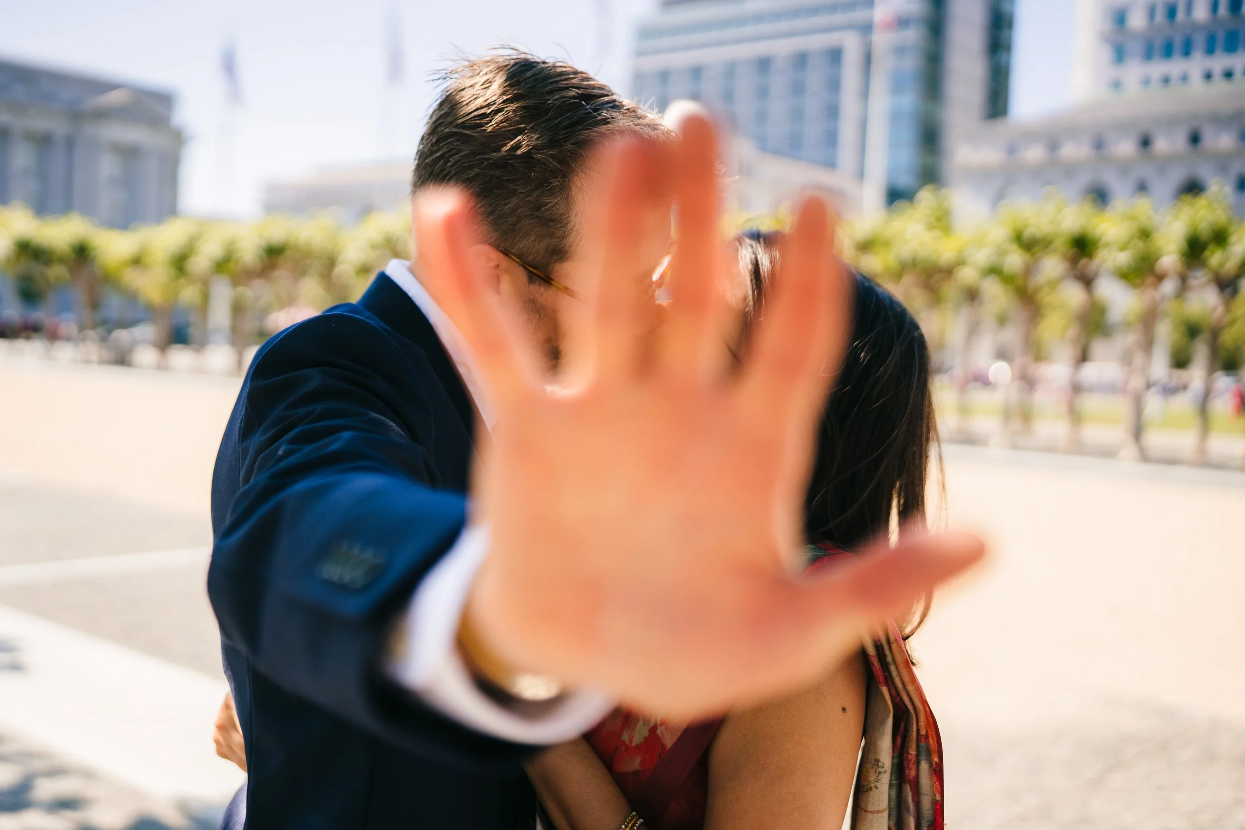 A close-up of a man and woman sharing an intimate moment outdoors, with the man's hand reaching out in front of the camera, obscuring their faces. The background shows city buildings and trees, indicating an urban park setting.