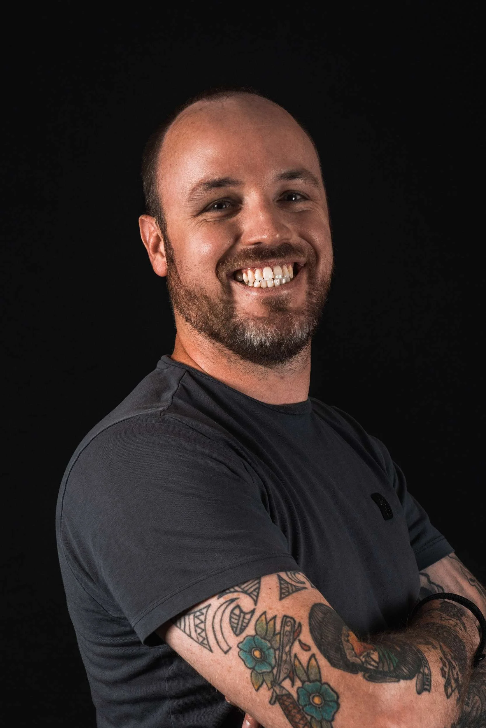A smiling man with tattoos on his arms, wearing a dark gray shirt, crossing his arms against a black background.