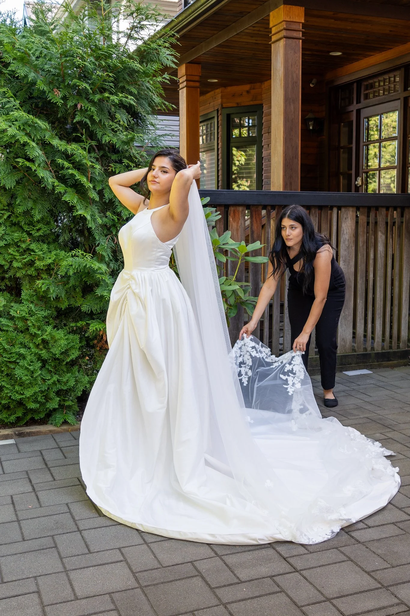 A beautiful bride in a white wedding gown stands outdoors while Shay from Shay Does Weddings adjusts the train of her dress.