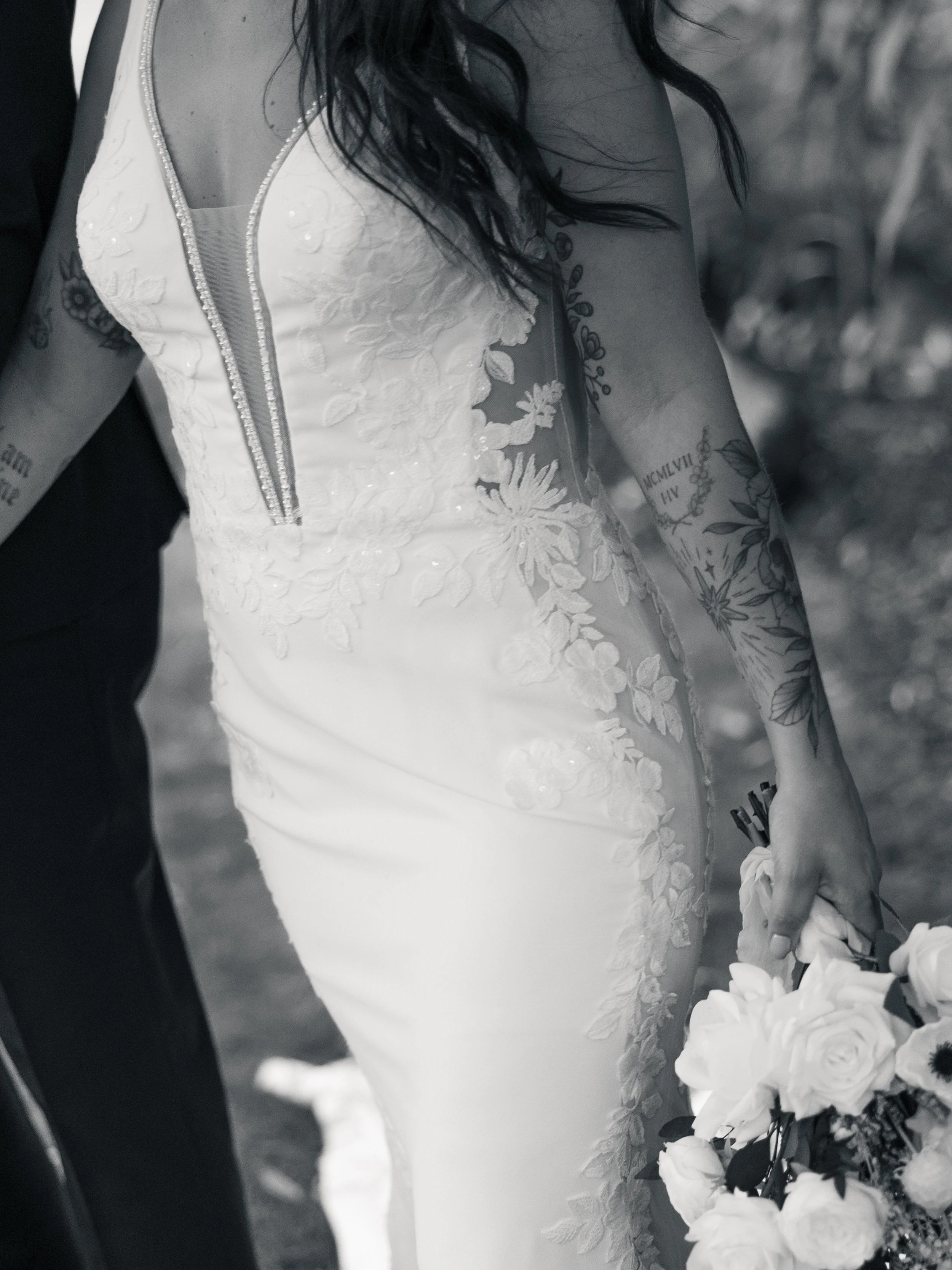 Close-up of a bride in a white wedding dress holding a bouquet of white roses and other flowers, displaying tattoos on her arm. Planned by Shay from Shay does Weddings.