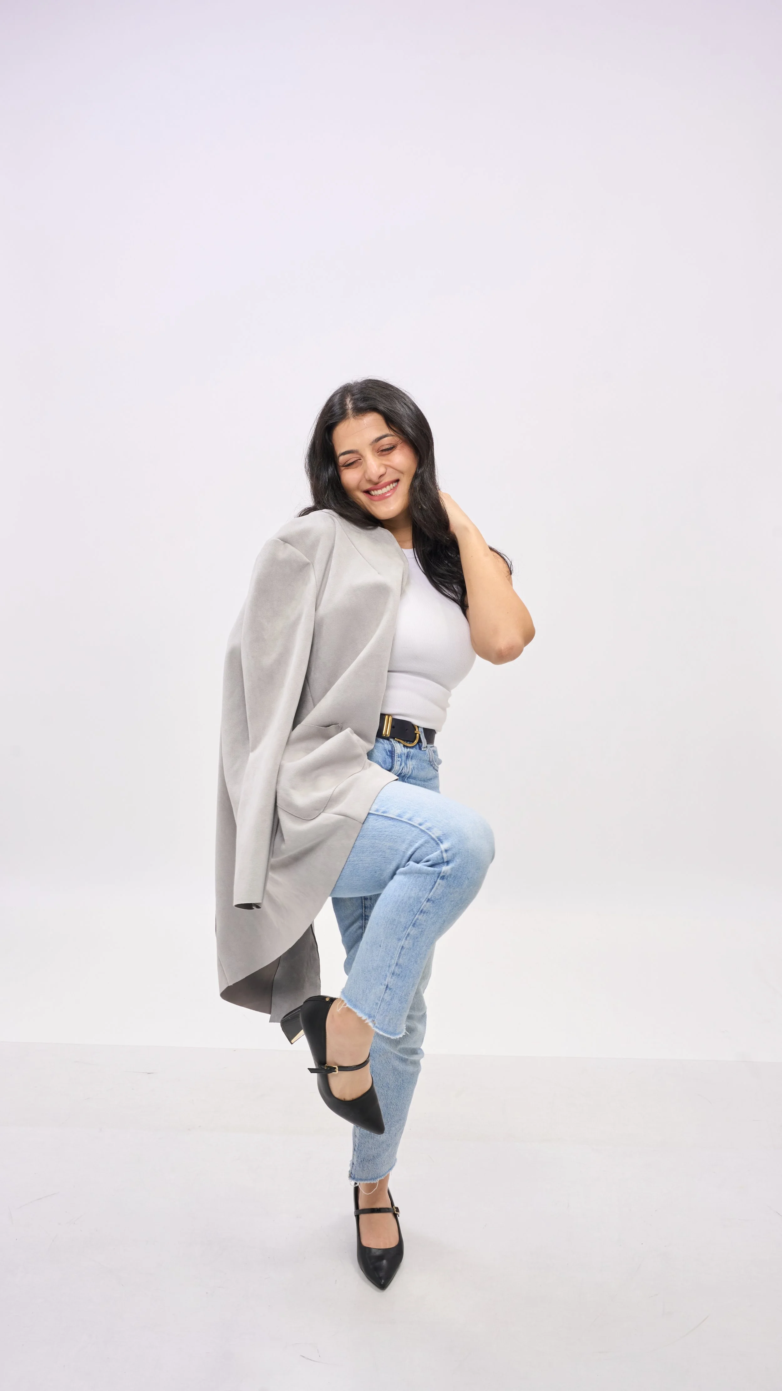 Shay from Shay does Weddings, wearing a grey blazer, white top, light blue jeans, and black heels, smiling and posing with one leg raised against a plain white background.