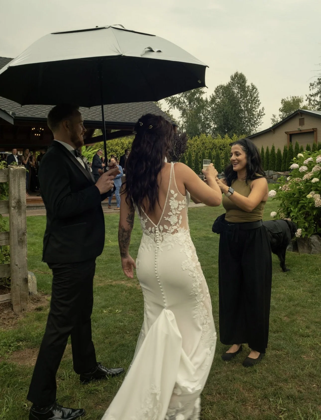A wedding scene outdoors with a bride, a groom in a black suit, and Shay from Shay Does Weddings. The groom is holding an umbrella over the bride as they share a moment, with guests in the background near a building and garden.