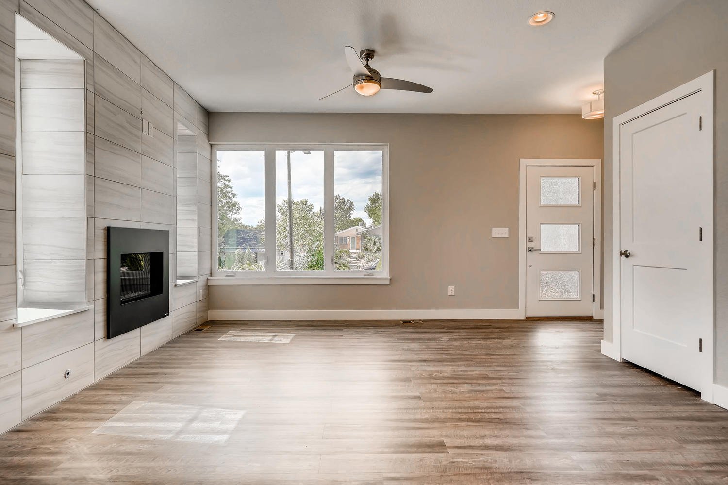 Empty living room with wood flooring, large window, white door, and wall-mounted fireplace