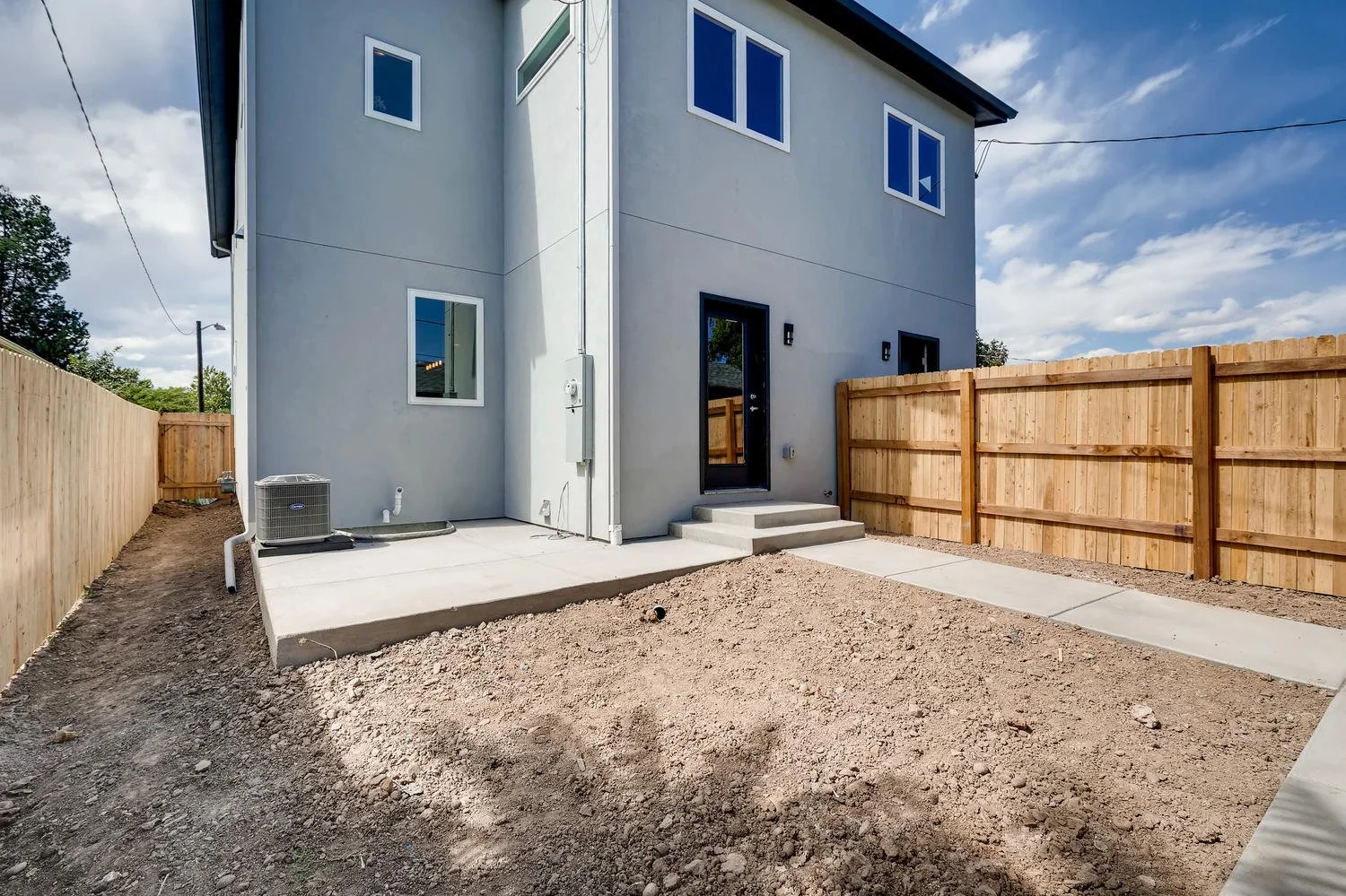 Backyard of a new two-story house with a concrete patio, dirt yard, wooden fence, and a pathway leading to the door. The house has a gray exterior with multiple windows, and an air conditioning unit is placed on the patio.