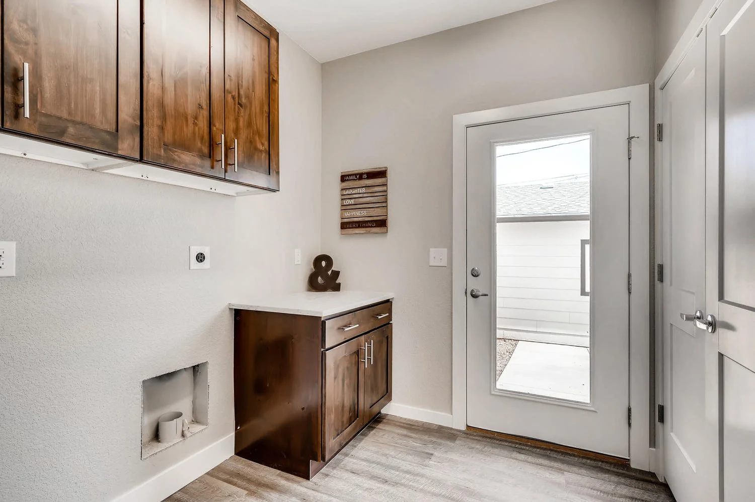 A laundry room with white walls and light wood flooring. There are dark wooden cabinets above and below a small counter. A decorative ampersand sits on the counter. To the right, there's a white door with a large glass panel leading outside. On the wall to the left of the door, there's a small wooden sign with phrases, and a small picture frame.