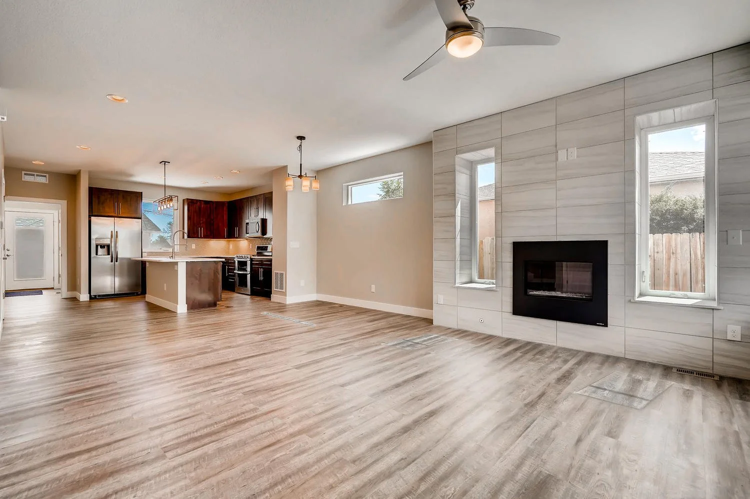 Open-concept living room and kitchen in a modern home with light wood flooring, a fireplace with a tiled wall, large windows, and a ceiling fan.